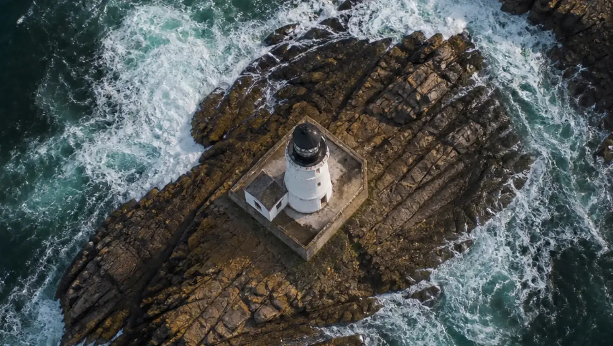 An aerial view of a white lighthouse on a rocky island, surrounded by crashing waves