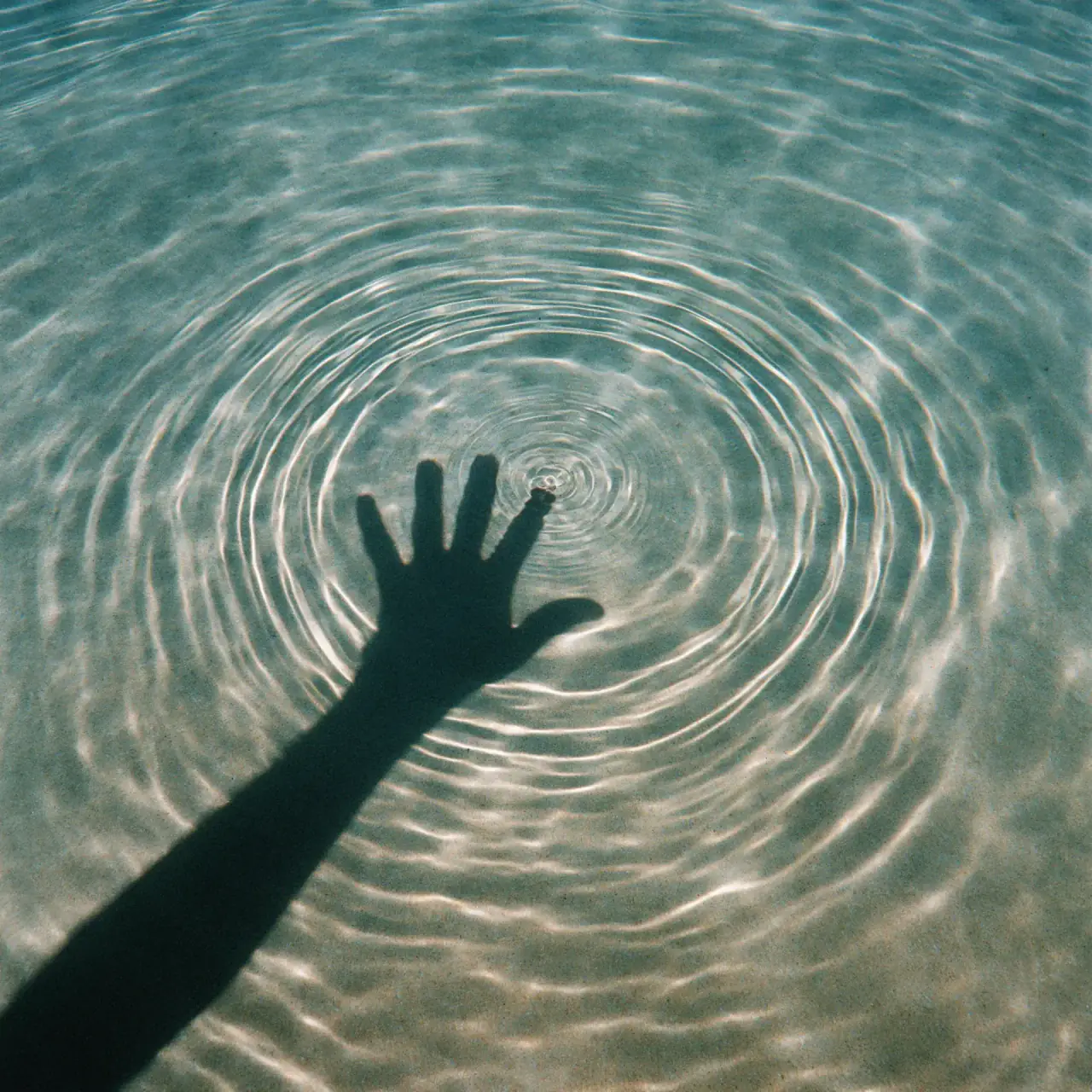 A hand's shadow reaching toward the center of concentric ripples on clear, sunlit water