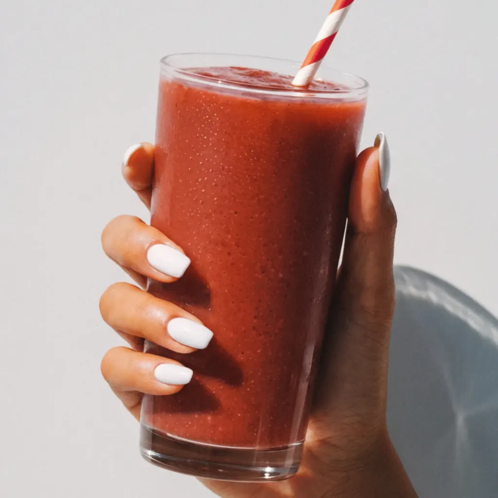 A hand with white nails holding a glass of red smoothie with a striped straw